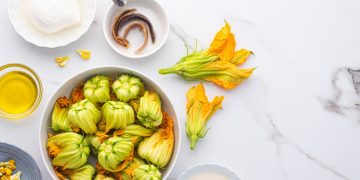 ricette con fiori di zucca su piano da cucina contenitore con fiori di zucchina, un bicchiere di olio EVO, in un piattino 3 acciughe e sulla destra un altro piattino con una mozzarella per illustrare i tanti modi su come cucinare i fiori di zucca