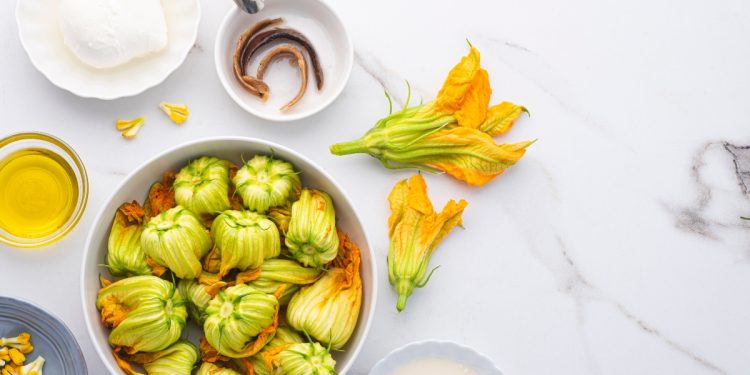 ricette con fiori di zucca su piano da cucina contenitore con fiori di zucchina, un bicchiere di olio EVO, in un piattino 3 acciughe e sulla destra un altro piattino con una mozzarella per illustrare i tanti modi su come cucinare i fiori di zucca