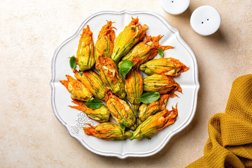 Fiori di zucca al forno: un delizioso contorno primaverile 3 su tavolo di legno beige un piatto bianco con fiori di zucca al forno. In alto a destra saliera e in basso a destra canovaccio colore mostarda