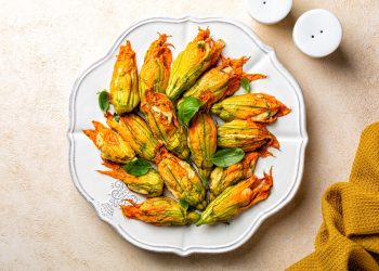 su tavolo di legno beige un piatto bianco con fiori di zucca al forno. In alto a destra saliera e in basso a destra canovaccio colore mostarda