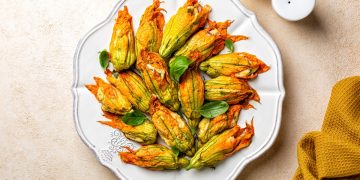 su tavolo di legno beige un piatto bianco con fiori di zucca al forno. In alto a destra saliera e in basso a destra canovaccio colore mostarda