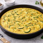 Frittata al forno: un secondo piatto vegetariano facile 1 In primo piano su un tavolo di marmo c'è una frittata al forno con delle zucchine. Sullo sfondo delle ciotoline bianco con pepe, Parmigiano.
