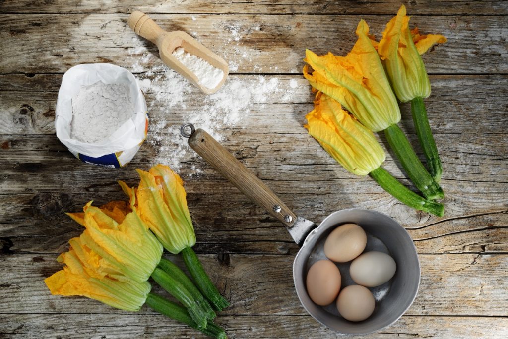 ricette con fiori di zucca: su tavolo di legno due mazzi di zucchine con fiori, un pentolino con 4 uova dentro, una ciotola di farina
