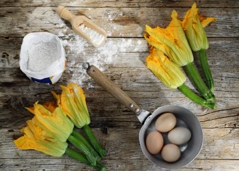 ricette con fiori di zucca: su tavolo di legno due mazzi di zucchine con fiori, un pentolino con 4 uova dentro, una ciotola di farina