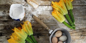 ricette con fiori di zucca: su tavolo di legno due mazzi di zucchine con fiori, un pentolino con 4 uova dentro, una ciotola di farina