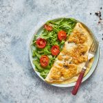 Frittata di albumi: una ricetta semplice e leggera 1 In primo piano su un tavolo di marmo grigio c'è un piatto fondo con una frittata di albumi e della rucola con i pomodorini, sul piatto c'è una forchetta con il manico in legno.