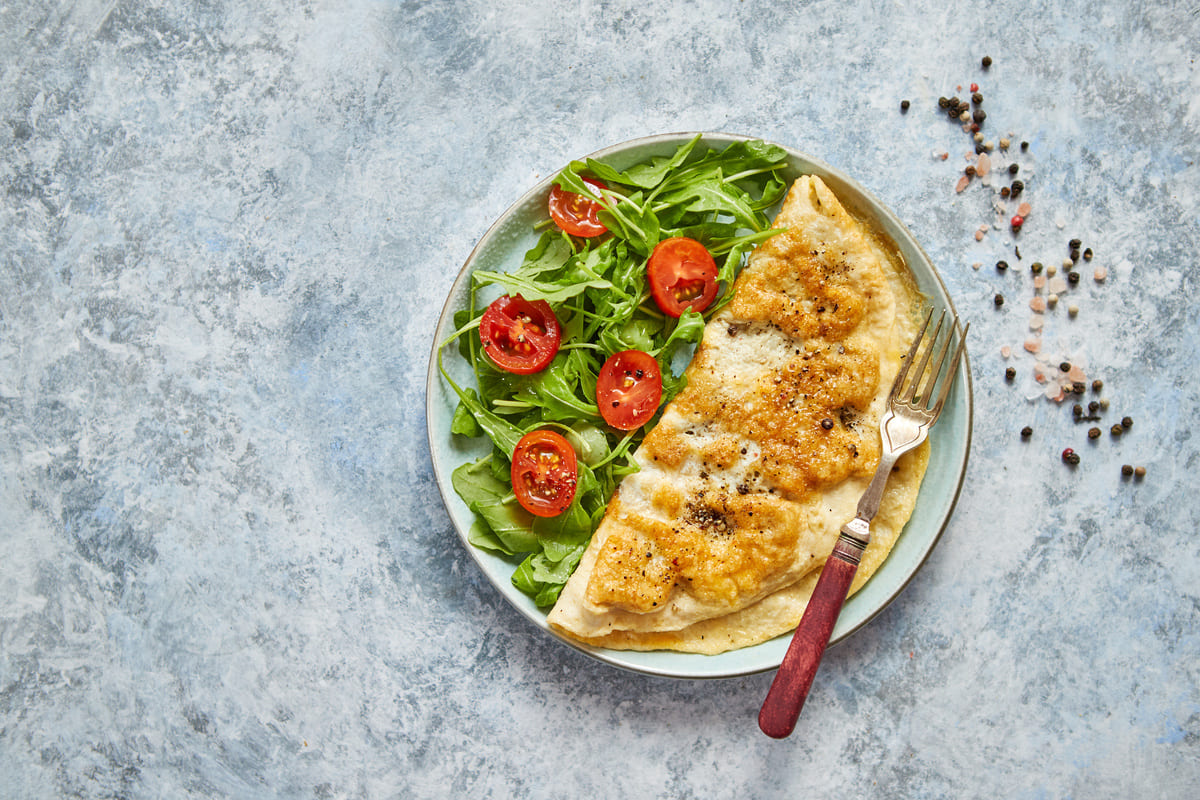 In primo piano su un tavolo di marmo grigio c'è un piatto fondo con una frittata di albumi e della rucola con i pomodorini, sul piatto c'è una forchetta con il manico in legno.