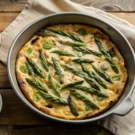 su tavolo di legno canovaccio beige con piatto da forno che contiene una frittata di asparagi ,sulla destra piattino con asparagi puliti
