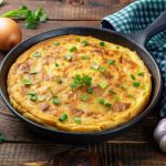 ricette con frittata: su tavolo di legno, teglia con frittata di patate e cipolle con attorno, cipolle bianche e rosse, un canovaccio verde a quadretti bianchi