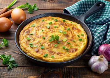 ricette con frittata: su tavolo di legno, teglia con frittata di patate e cipolle con attorno, cipolle bianche e rosse, un canovaccio verde a quadretti bianchi