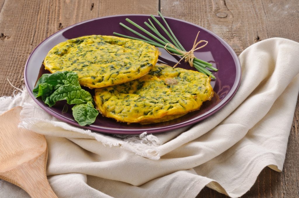 ricette con frittata: su tavolo di legno, canovaccio beige con sopra un piatto viola con frittata di spinaci, erba cipolline e una foglia di spinaci freschi