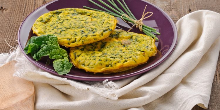 ricette con frittata: su tavolo di legno, canovaccio beige con sopra un piatto viola con frittata di spinaci, erba cipolline e una foglia di spinaci freschi