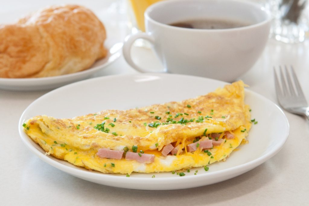 Omelette prosciutto e formaggio 1 su un tavolo da colazione in primo piano piatto bianco con omelette prosciutto e formaggio, dietro una tazza di caffè e accanto una brioche in un piatto bianco