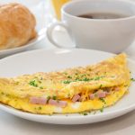 su un tavolo da colazione in primo piano piatto bianco con omelette prosciutto e formaggio, dietro una tazza di caffè e accanto una brioche in un piatto bianco