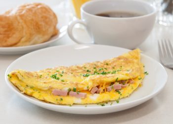 su un tavolo da colazione in primo piano piatto bianco con omelette prosciutto e formaggio, dietro una tazza di caffè e accanto una brioche in un piatto bianco