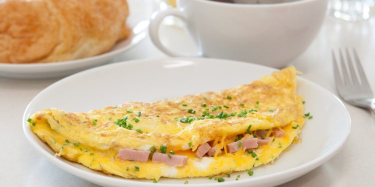 su un tavolo da colazione in primo piano piatto bianco con omelette prosciutto e formaggio, dietro una tazza di caffè e accanto una brioche in un piatto bianco