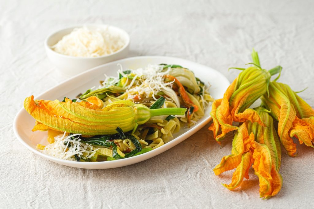 ricette con fiori di zucca: su tavolo di legno bianco, piatto piccolo con la pasta e sopra un fiore di zucca. Accanto 3 fiori e dietro una ciotola con parmigiano grattugiato