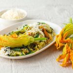 ricette con fiori di zucca: su tavolo di legno bianco, piatto piccolo con la pasta e sopra un fiore di zucca. Accanto 3 fiori e dietro una ciotola con parmigiano grattugiato