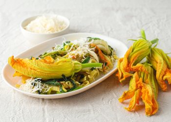 ricette con fiori di zucca: su tavolo di legno bianco, piatto piccolo con la pasta e sopra un fiore di zucca. Accanto 3 fiori e dietro una ciotola con parmigiano grattugiato