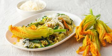 ricette con fiori di zucca: su tavolo di legno bianco, piatto piccolo con la pasta e sopra un fiore di zucca. Accanto 3 fiori e dietro una ciotola con parmigiano grattugiato
