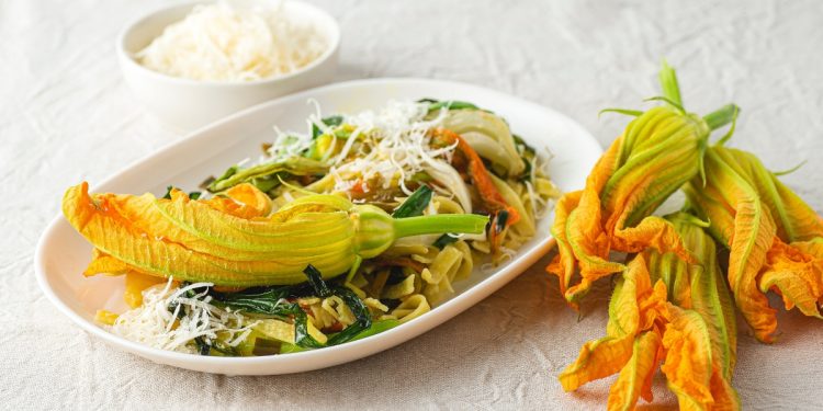 ricette con fiori di zucca: su tavolo di legno bianco, piatto piccolo con la pasta e sopra un fiore di zucca. Accanto 3 fiori e dietro una ciotola con parmigiano grattugiato