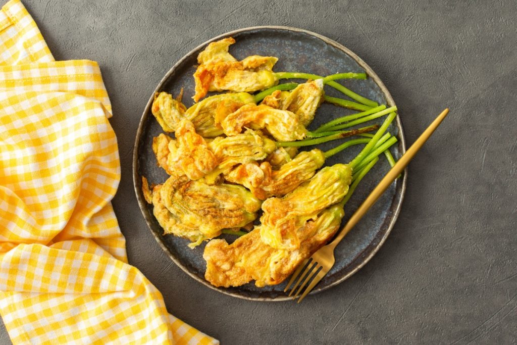Pastella per fiori di zucca senza uovo e senza lievito 5 pastella per fiori di zucca su alcuni fiori di zucca fritti presentati su un piatto tondo scuro, un tovagliolo a quadretti bianchi e gialli accanto