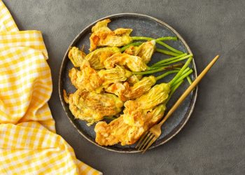pastella per fiori di zucca su alcuni fiori di zucca fritti presentati su un piatto tondo scuro, un tovagliolo a quadretti bianchi e gialli accanto