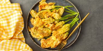 pastella per fiori di zucca su alcuni fiori di zucca fritti presentati su un piatto tondo scuro, un tovagliolo a quadretti bianchi e gialli accanto