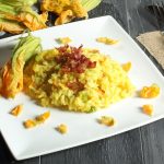 su tavolo di legno scuro, piatto bianco quadrato con risotto con fiori di zucca e all'angolo fiore di zucca per decorare, anche dietro e sulla destra forchetta e coltello