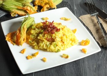 su tavolo di legno scuro, piatto bianco quadrato con risotto con fiori di zucca e all'angolo fiore di zucca per decorare, anche dietro e sulla destra forchetta e coltello