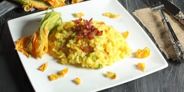 su tavolo di legno scuro, piatto bianco quadrato con risotto con fiori di zucca e all'angolo fiore di zucca per decorare, anche dietro e sulla destra forchetta e coltello