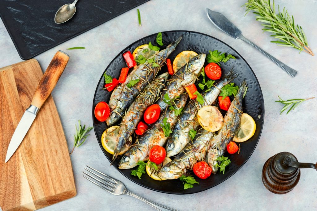 su piano cottura, piatto nero con sardine al forno con pomodorini e limoni a fette, a destra tagliere con coltello e sulla sinistra rosmarino