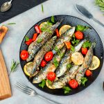 su piano cottura, piatto nero con sardine al forno con pomodorini e limoni a fette, a destra tagliere con coltello e sulla sinistra rosmarino