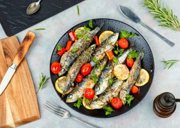 su piano cottura, piatto nero con sardine al forno con pomodorini e limoni a fette, a destra tagliere con coltello e sulla sinistra rosmarino