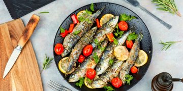 su piano cottura, piatto nero con sardine al forno con pomodorini e limoni a fette, a destra tagliere con coltello e sulla sinistra rosmarino