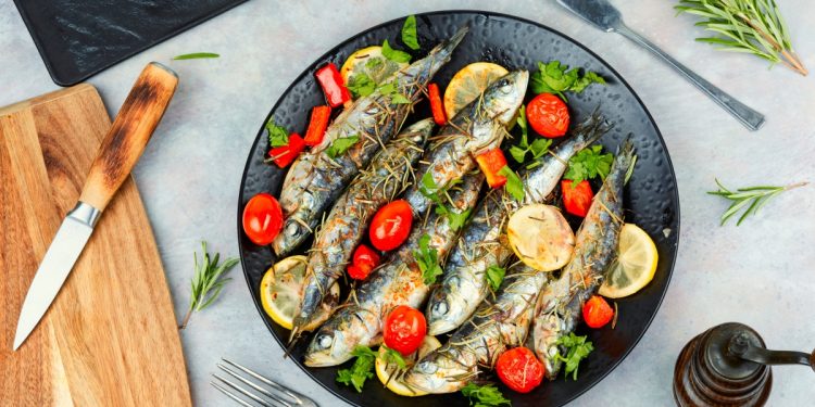 su piano cottura, piatto nero con sardine al forno con pomodorini e limoni a fette, a destra tagliere con coltello e sulla sinistra rosmarino