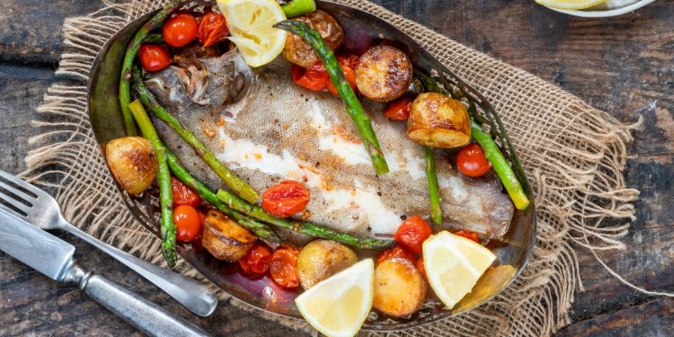 su tavolo di legno sotto tavolo stile bohémien con un piatto con sogliola al forno, patate, asparagi, pomodorini con alcune fette di limoni . Sulla sinistra forchetta e coltello