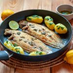 Sogliola alla mugnaia: un secondo sano pronto in 10 minuti 1 In primo piano su un tavolo di legno, in una padella c'è una sogliola alla mugnaia con delle patate novelle condite con limone e prezzemolo. Sul tavolo si trovano dei limoni gialli, un lime e del prezzemolo in una piccola ciotola.