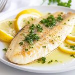 su tavolo, primo piano di un piatto bianco con una sogliola alla mugnaia con prezzemolo e fette di limone