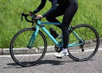 Bici da corsa verde acqua in primo piano su sfondo una strada, con telaio aerodinamico e dettagli tecnici visibili