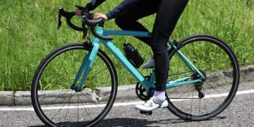 Bici da corsa verde acqua in primo piano su sfondo una strada, con telaio aerodinamico e dettagli tecnici visibili