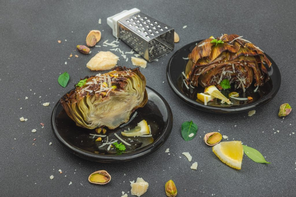 su tavolo di marmo, 2 piatti neri con carciofi bolliti, attorno pistacchi, fette di limone, grattugia e parmigiano reggiano