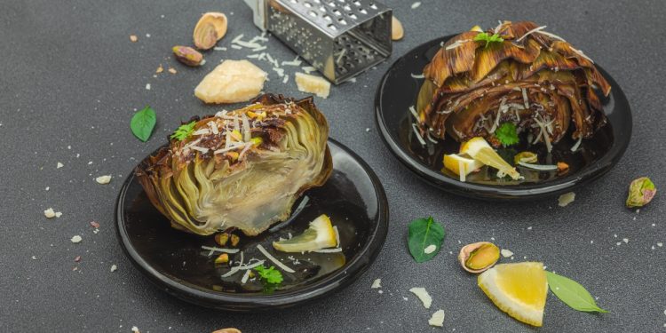 su tavolo di marmo, 2 piatti neri con carciofi bolliti, attorno pistacchi, fette di limone, grattugia e parmigiano reggiano