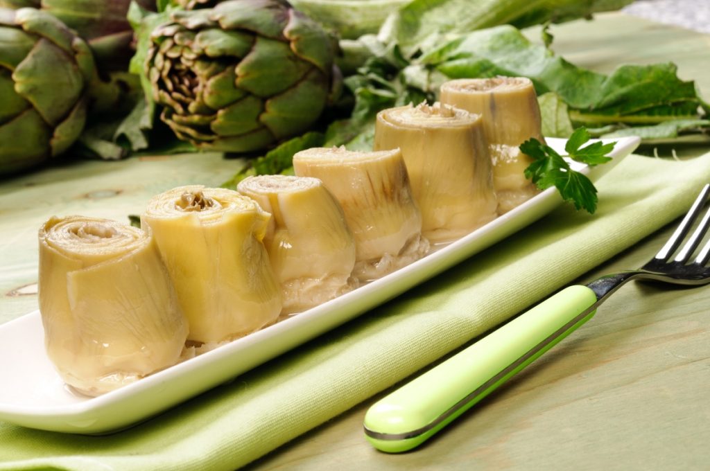 su tavolo da cucina piatto rettangolare verde sopra una tovagliolo verde con 6 carciofini sott'olio e sulla destra una forchetta verde. dietro il piatto carciofi freschi