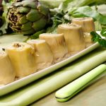 su tavolo da cucina piatto rettangolare verde sopra una tovagliolo verde con 6 carciofini sott'olio e sulla destra una forchetta verde. dietro il piatto carciofi freschi