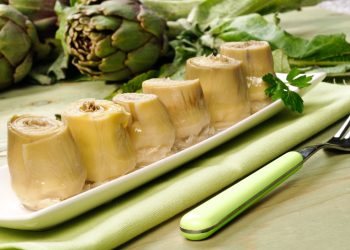 su tavolo da cucina piatto rettangolare verde sopra una tovagliolo verde con 6 carciofini sott'olio e sulla destra una forchetta verde. dietro il piatto carciofi freschi