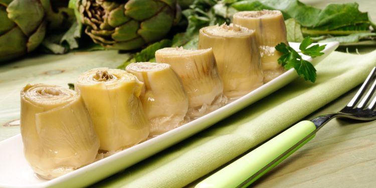 su tavolo da cucina piatto rettangolare verde sopra una tovagliolo verde con 6 carciofini sott'olio e sulla destra una forchetta verde. dietro il piatto carciofi freschi