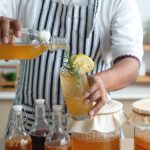 come fare kombucha: uomo in cucina con grembiule che sta preparando kombucha in casa e riempie i bicchieri