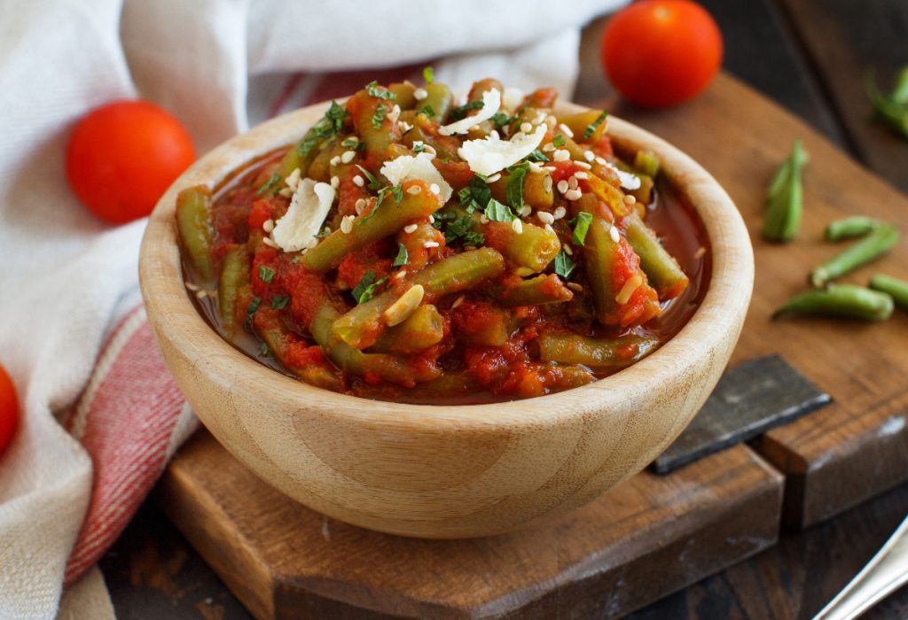 Fagiolini all'uccelletto: la ricetta toscana tradizionale 4 su tavolo, tagliere con ciotola in marmo con fagiolini all'uccelletto con alcuni fagiolini lavati e tagliati e dietro pomodorini