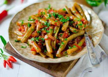 su tavolo apparecchiato tagliere con sopra un piatto con fagiolini in umido con pomodorini e prezzemolo e una forchetta sulla destra peperoncini